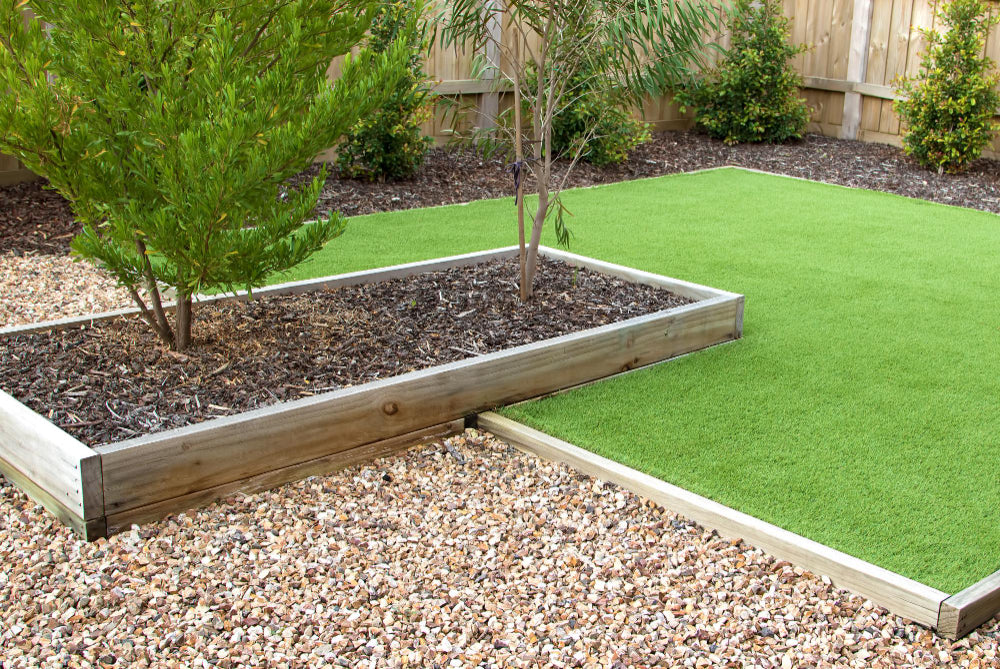 A landscaped garden with a bark border and central bed containing shrubs and trees, surrounded by a square of artificial lawn.